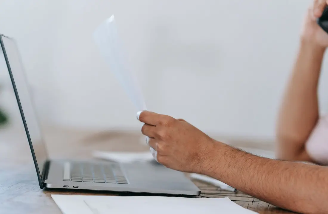 Crop anonymous male reading document and talking on mobile phone while sitting at desk with laptop