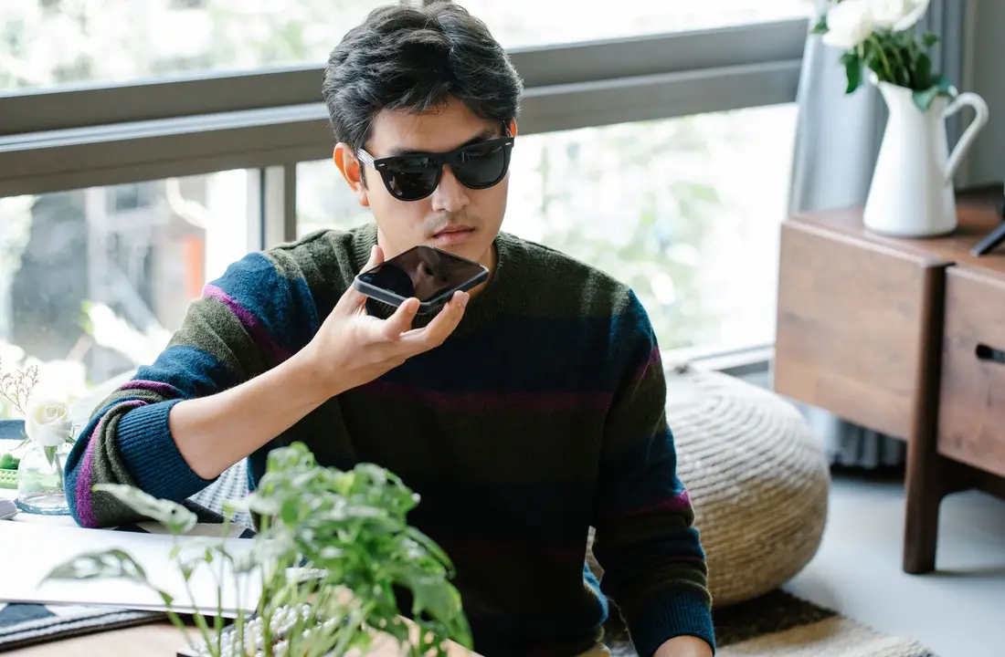 A man wearing sunglasses uses a smartphone's voice assistant in a cozy indoor setting.