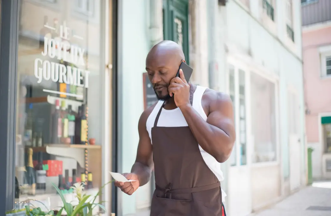 Man in apron making a business call outside a gourmet store in Portugal, checking notes.
