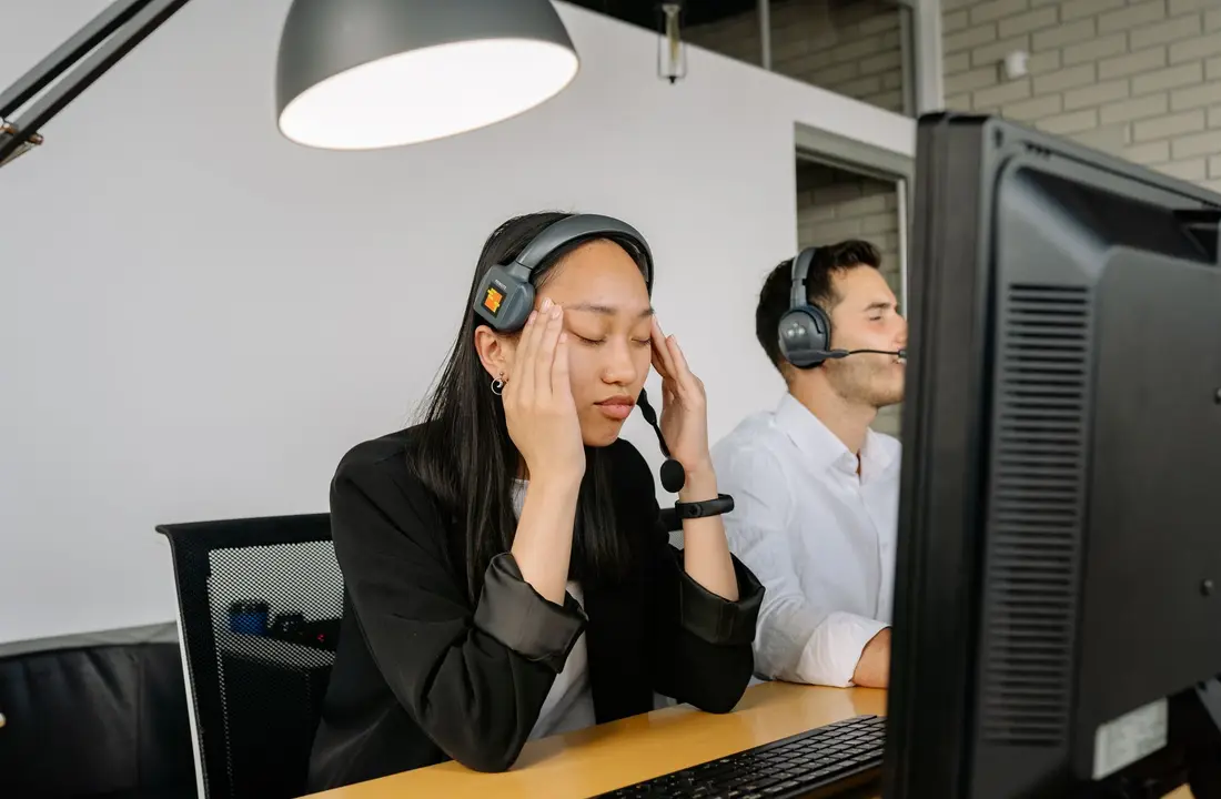 Asian woman at call center desk looking stressed, eyes closed, in office setting.