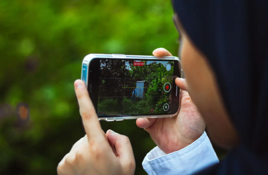 Close-up of person using smartphone to film a green outdoor scene in daylight.