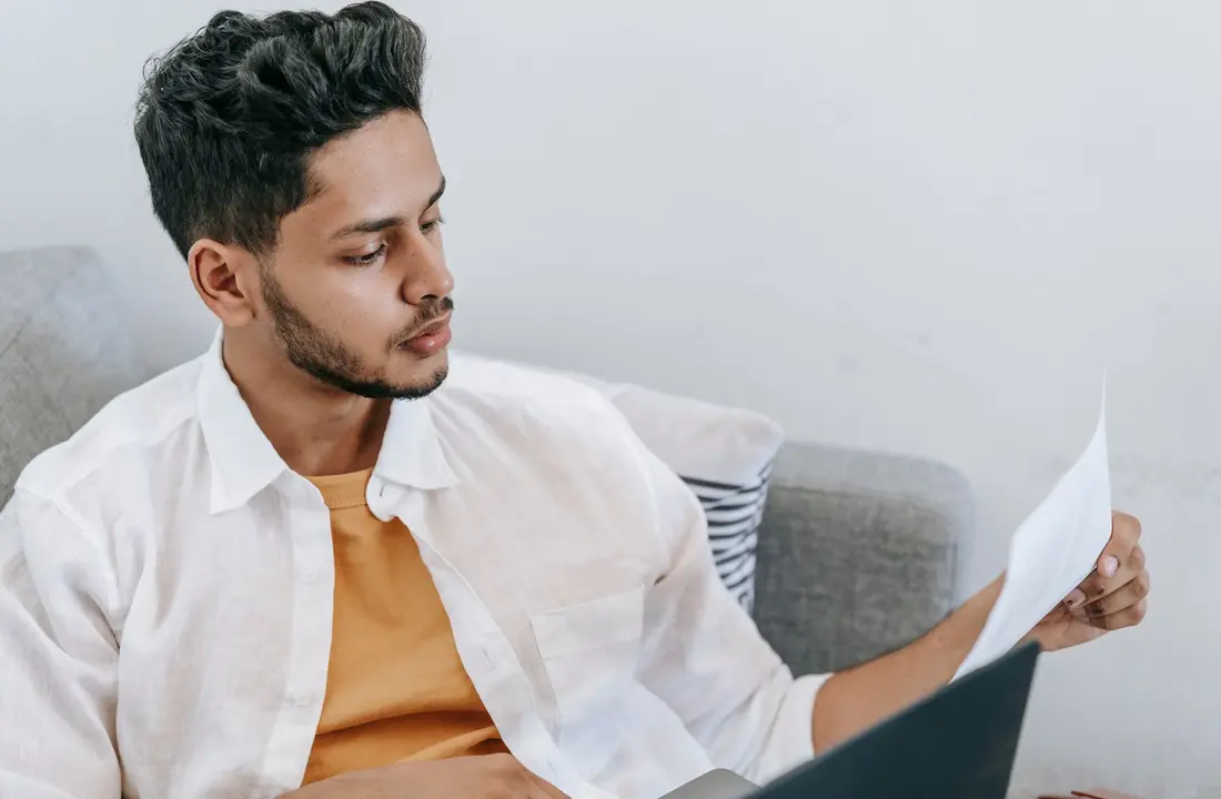 Concentrated young ethnic male wearing casual outfit reading document and browsing netbook while resting on cozy couch