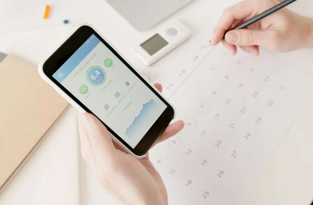 Close-up of hands using a smartphone to track health stats while planning on a calendar.